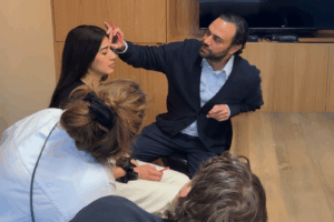 A medical professional (Dr. Moubayed) in a dark cardigan demonstrates a facial aesthetics technique, marking a treatment area for Botox, on a woman's forehead in front of a small group of students.